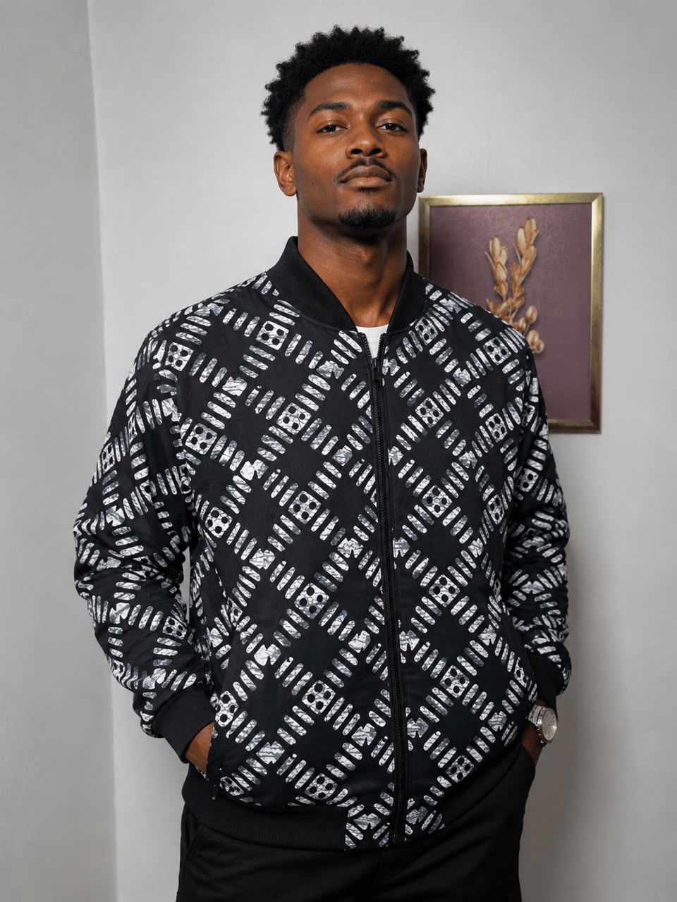 A man wearing a black and white patterned bomber jacket, standing against a light gray wall with a framed picture in the background.