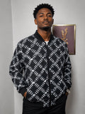 A man wearing a black and white patterned bomber jacket, standing against a light gray wall with a framed picture in the background.